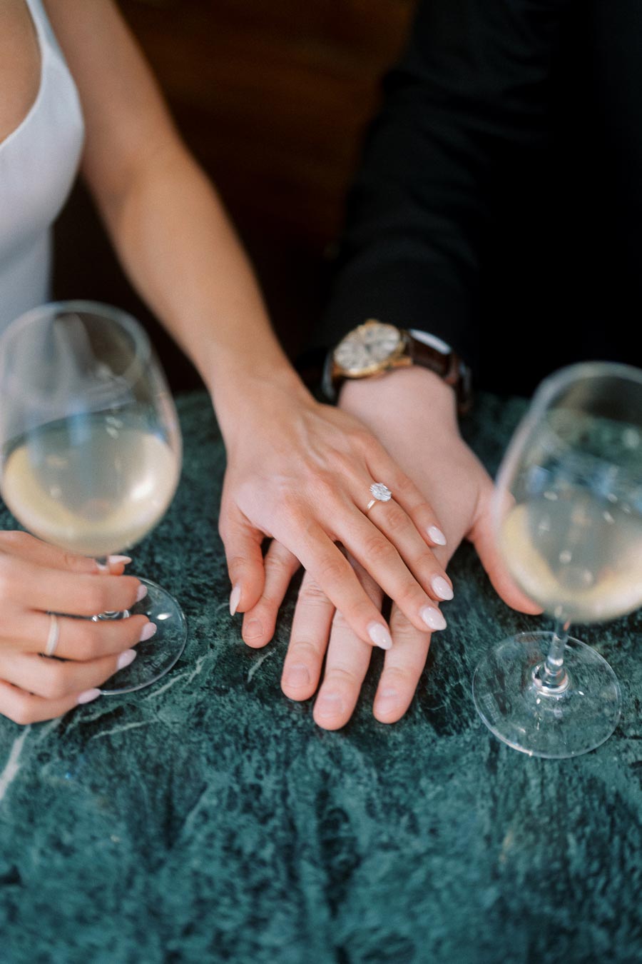 Close-up of a couple's hands with an engagement ring resting on a green marble table, two wine glasses in the foreground, romantic indoor scene.