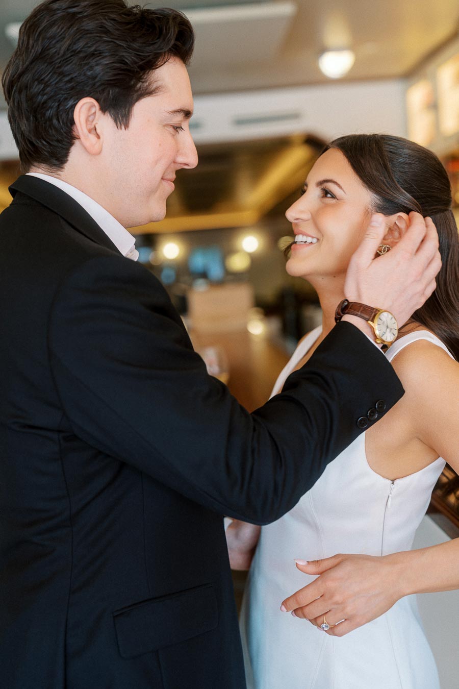 A couple dressed in formal attire shares a tender moment, smiling and gazing at each other inside a warmly lit bar in Ontario, Canada.