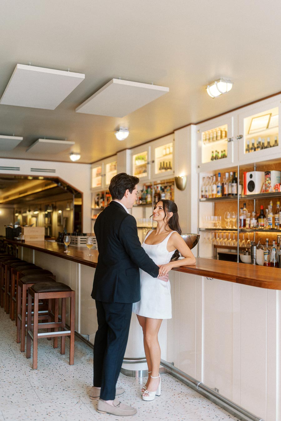 A couple stands together at a modern bar, smiling and gazing at each other lovingly, with shelves of bottles in the background.