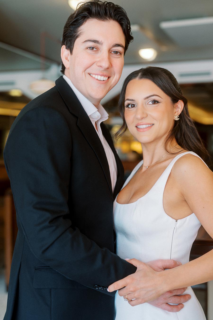 A smiling couple embraces indoors, dressed in formal attire, in a warmly lit Ontario bar setting during their engagement photoshoot.