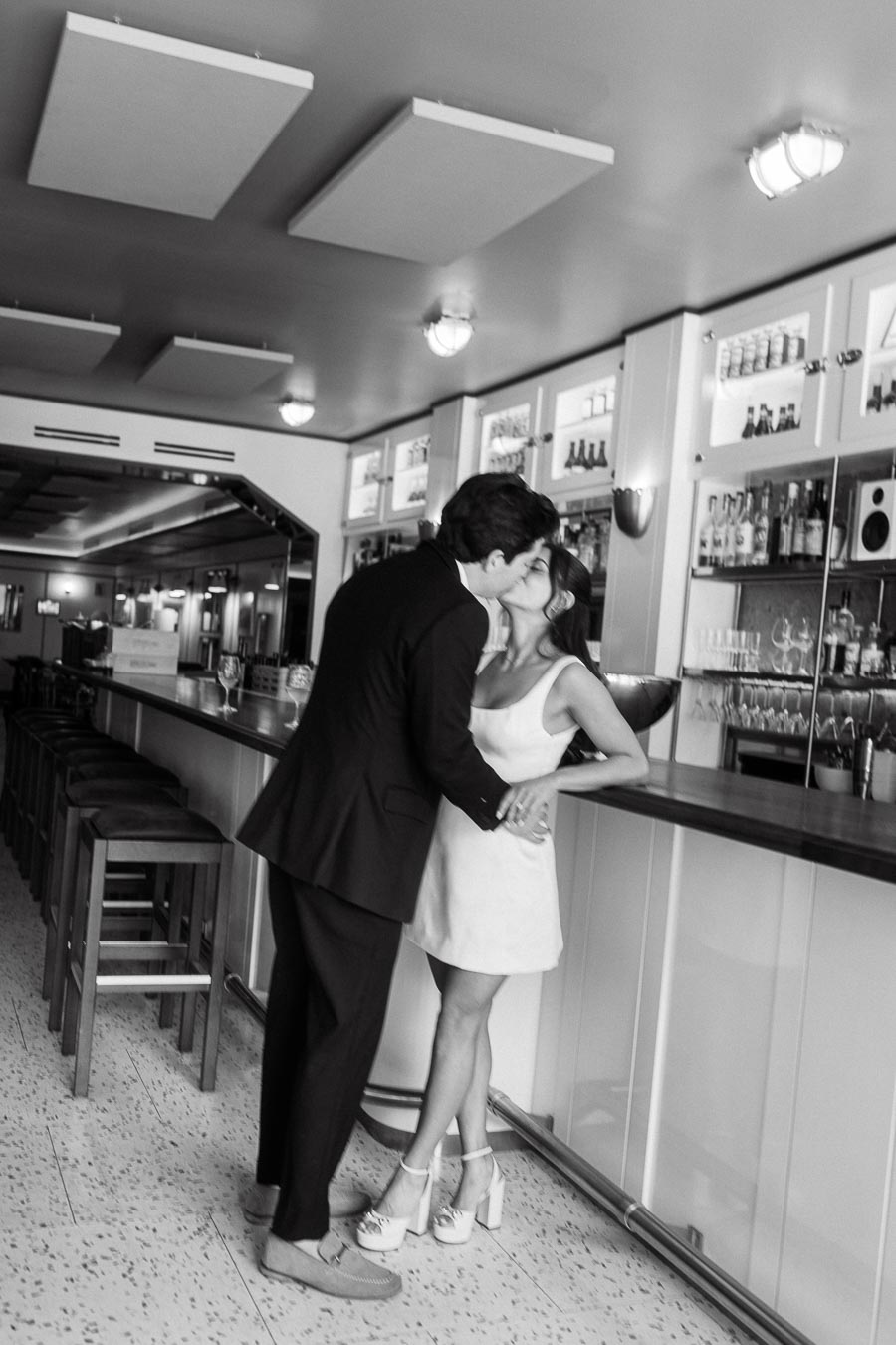 A couple in formal attire kisses affectionately beside a bar counter in a stylish indoor setting, Ontario, Canada, 2026.