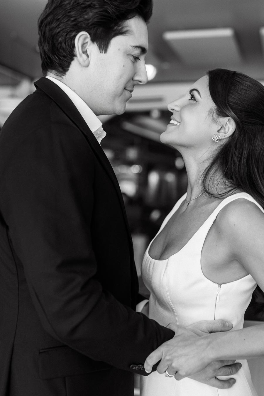 A black and white photo of a couple indoors, smiling at each other closely and holding hands in a romantic embrace.