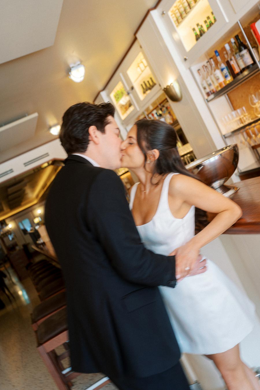 A couple dressed in formal attire shares a kiss while standing by the bar in a warmly lit, modern indoor bar setting.
