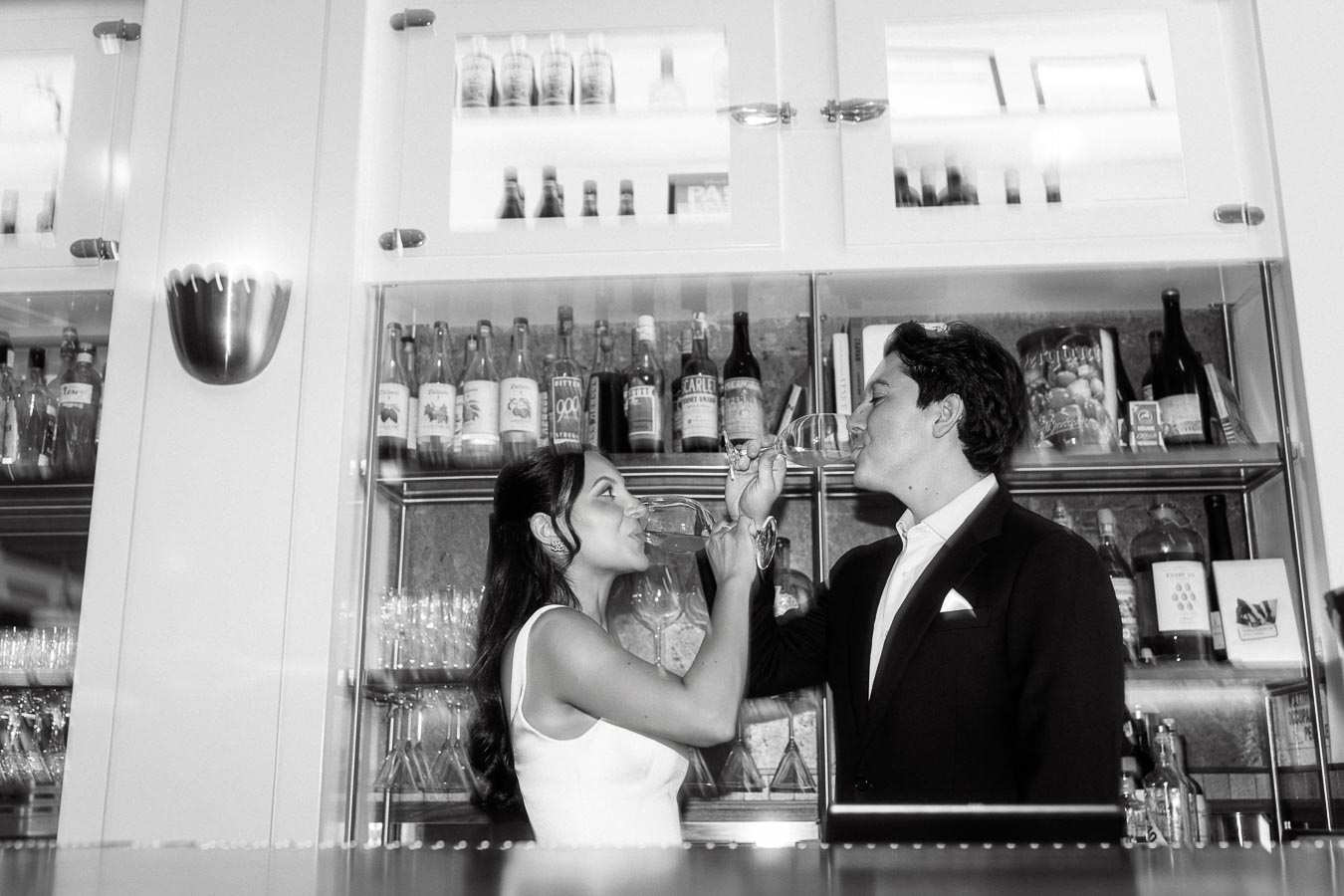 A couple dressed elegantly clinks wine glasses and smiles at each other in a stylish indoor bar, surrounded by bottles and glassware.