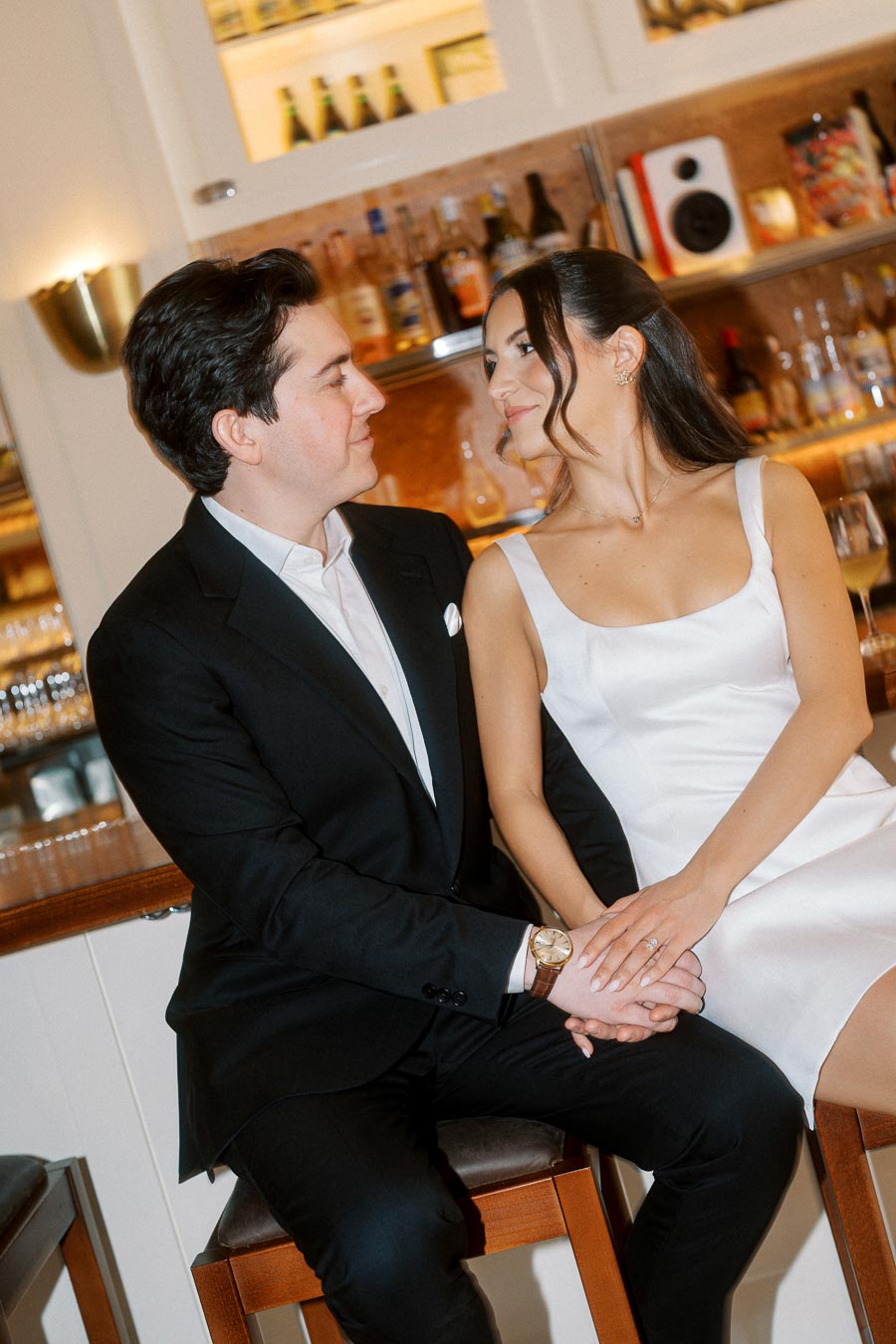 A couple dressed elegantly sits close together at a bar in Ontario, Canada, holding hands and gazing into each other's eyes with smiles.