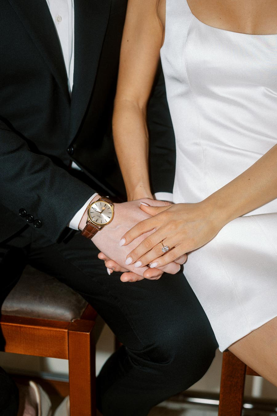 Close-up of an engaged couple sitting together, holding hands. The woman wears a white dress and engagement ring, the man a black suit and watch.