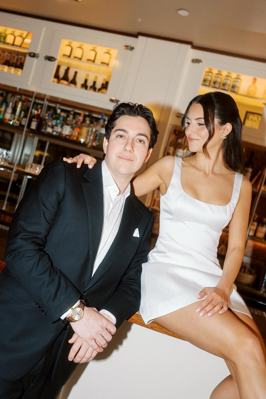 A couple in formal attire posing together at a bar, the woman sitting on the counter and smiling at the man, with bottles visible in the background.