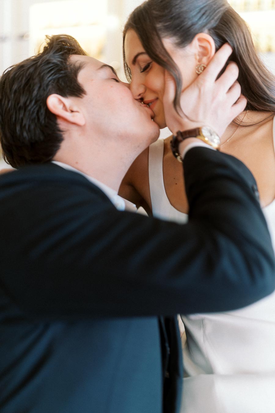 Close-up of a couple sharing a tender kiss, the man gently holding the woman's face, both dressed in formal attire, in a softly lit setting.