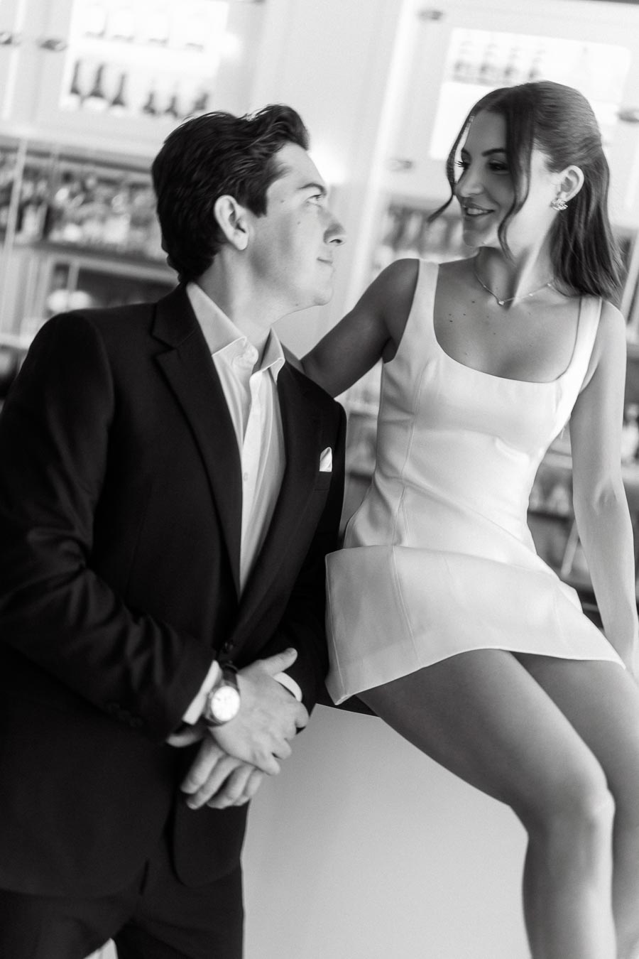 A couple dressed elegantly, the woman in a white dress sitting on a bar counter, smiling and looking at her partner in a suit, at a modern indoor bar.