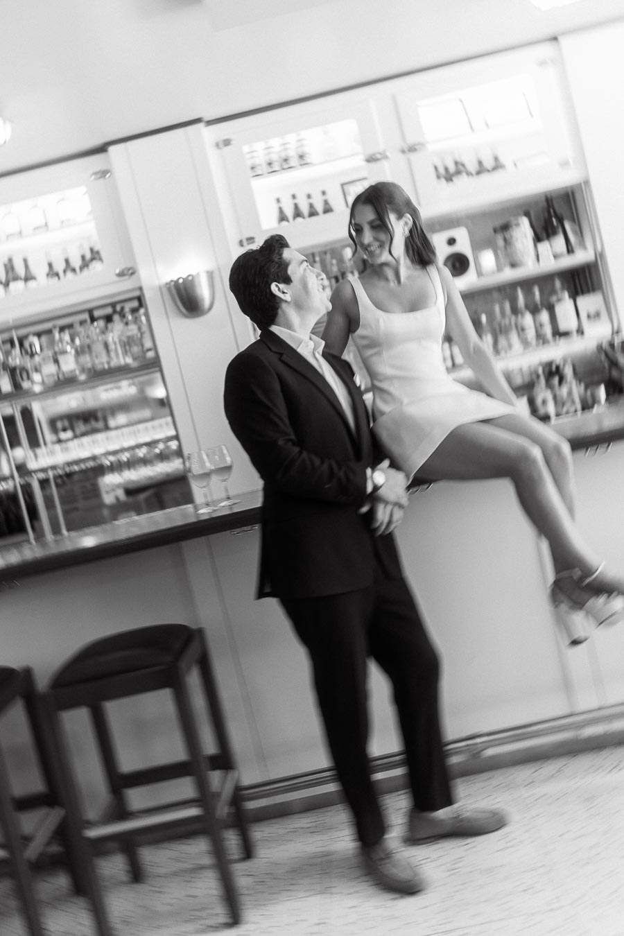 A couple in formal attire sharing a joyful moment at a modern bar, with the woman sitting on the counter and the man standing beside her, both smiling.