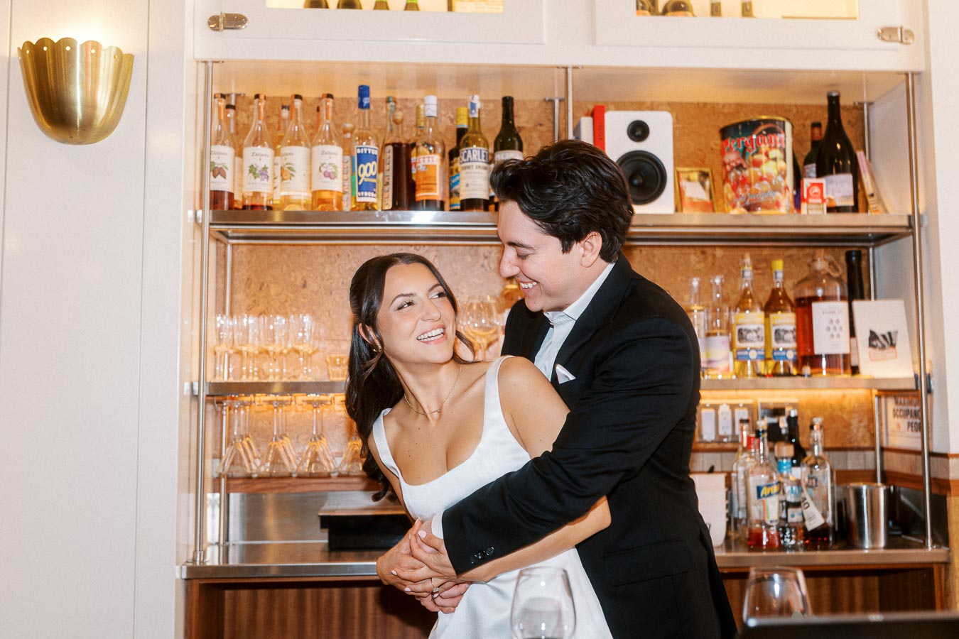 Smiling couple in formal attire embracing and laughing together in front of a bar shelf with bottles and glasses, Ontario, Canada.