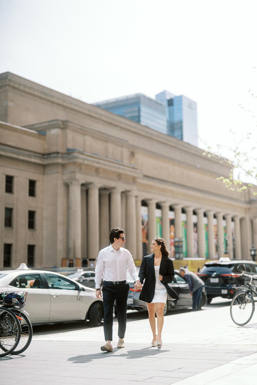 A couple holding hands and walking in front of Toronto Union Station with taxis and city buildings in the background.