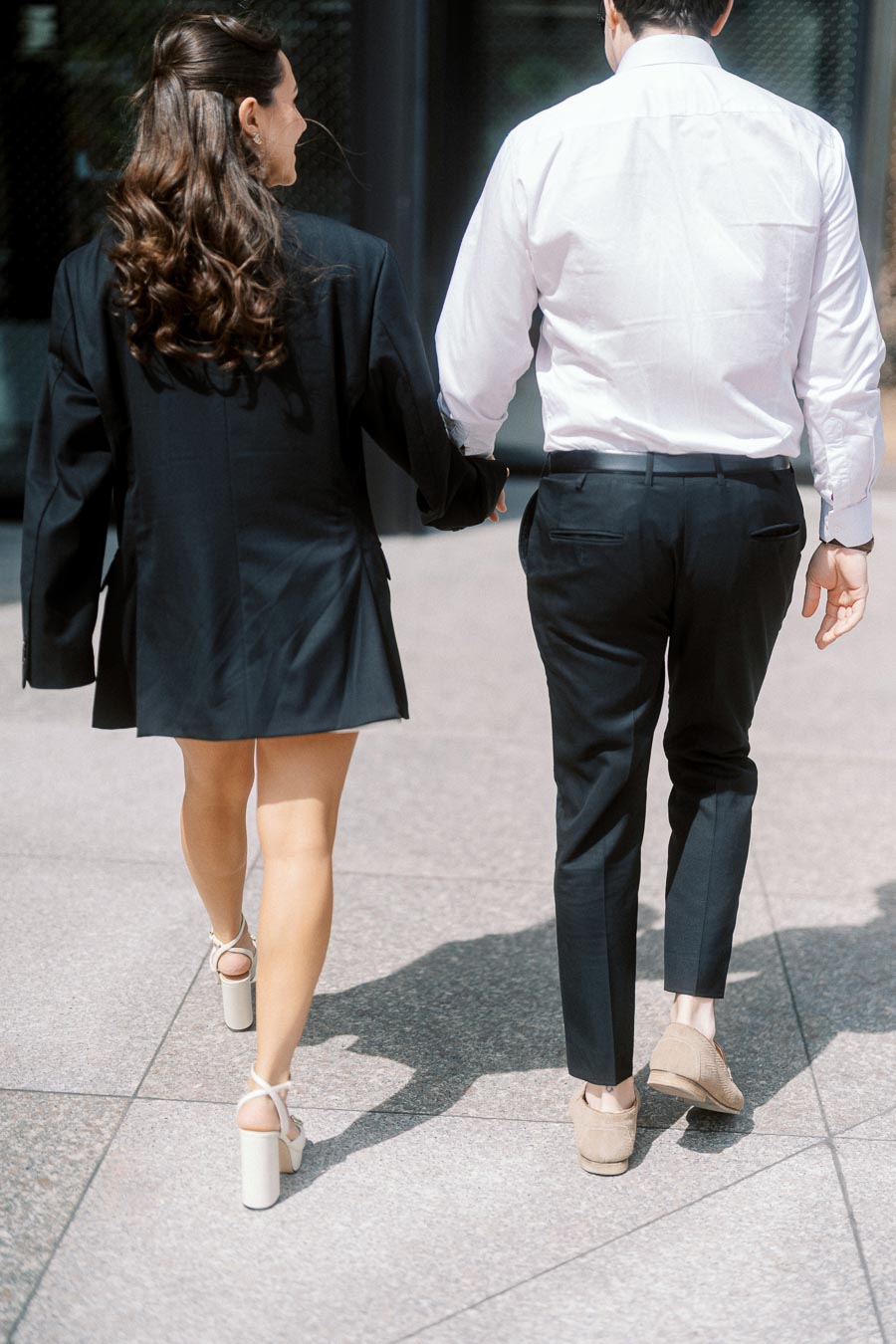 A couple walks hand in hand on a city sidewalk, dressed in formal attire, with the sun casting their shadows on the pavement.