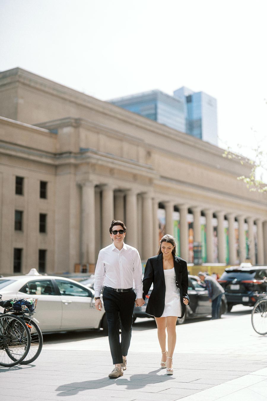 A couple holding hands and walking on a city sidewalk in front of Union Station Toronto, with cars and bicycles nearby in daylight.