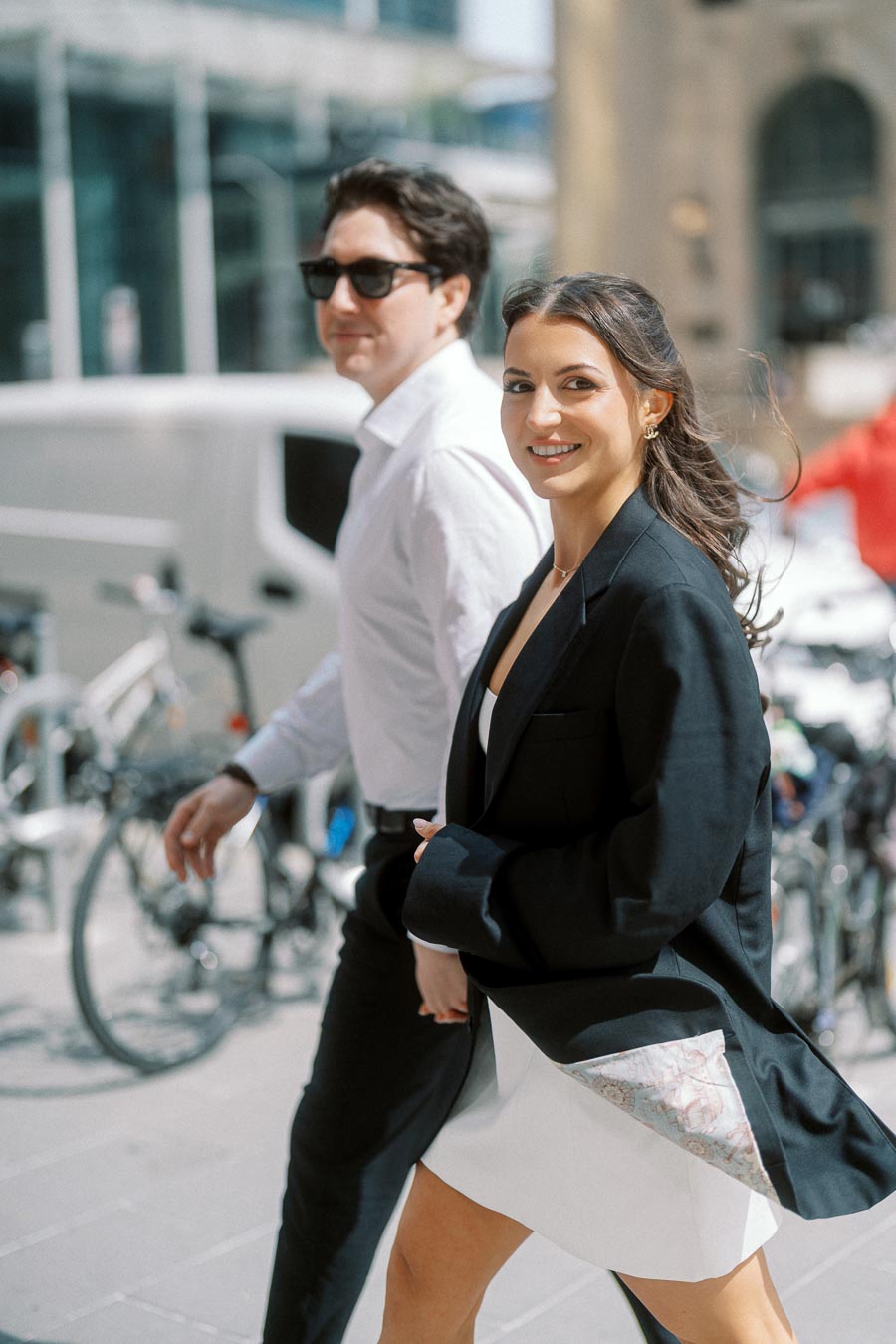A couple walking side by side outdoors in downtown Ontario, both smiling, with city bikes and urban buildings in the background.