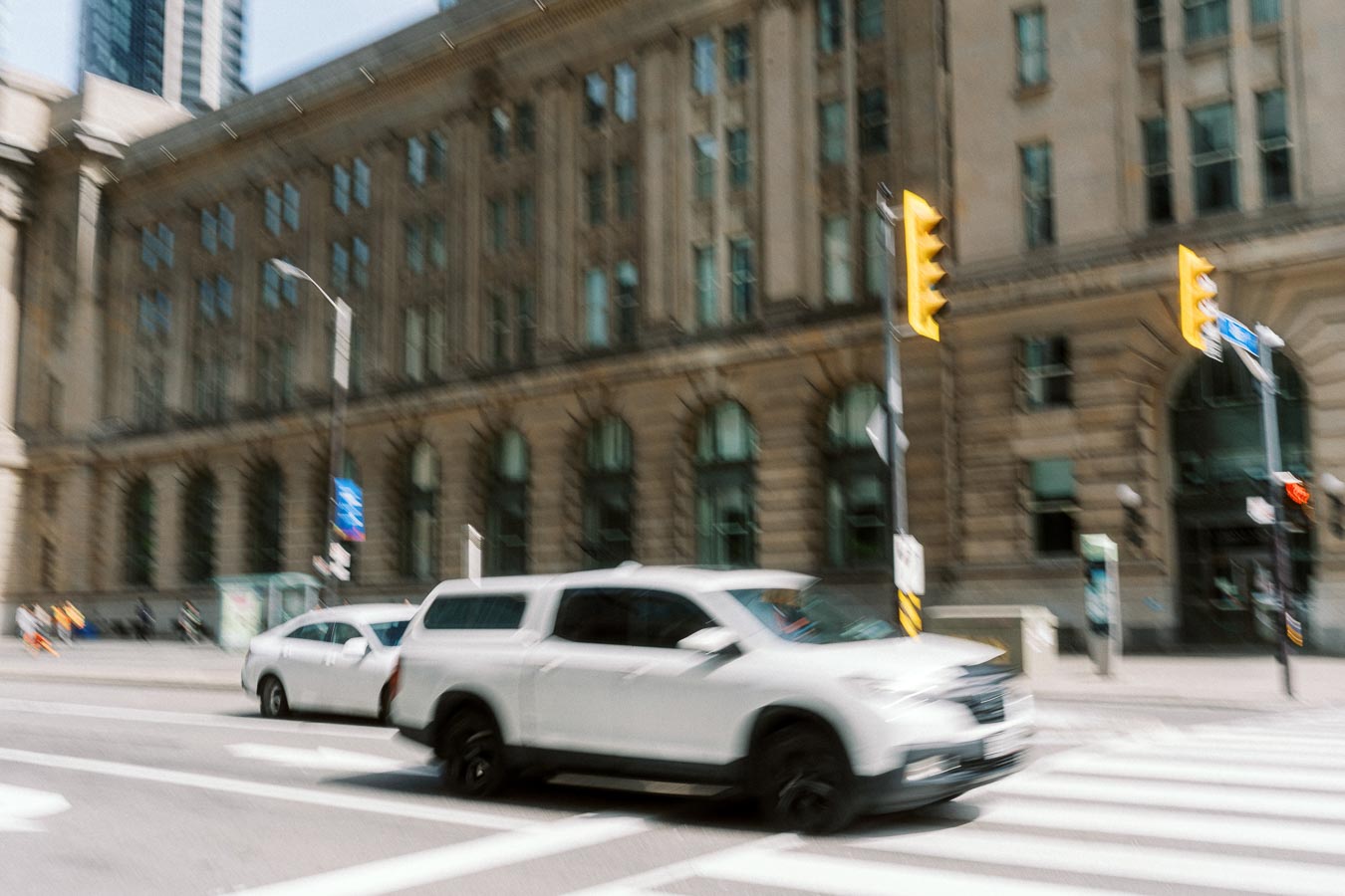 White SUV driving through a city crosswalk in front of a large historic stone building with traffic lights in downtown Toronto.