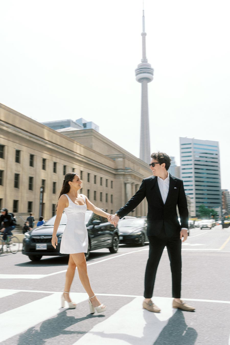 Couple in formal attire holding hands while crossing a city street near Union Station with the CN Tower visible in the background.