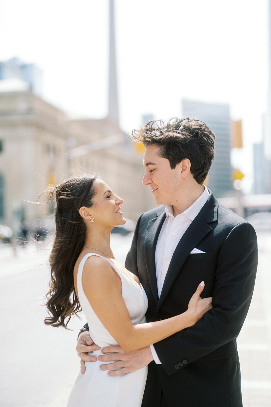A couple dressed in formal attire embraces and smiles at each other on a city street in downtown Toronto with urban buildings visible.