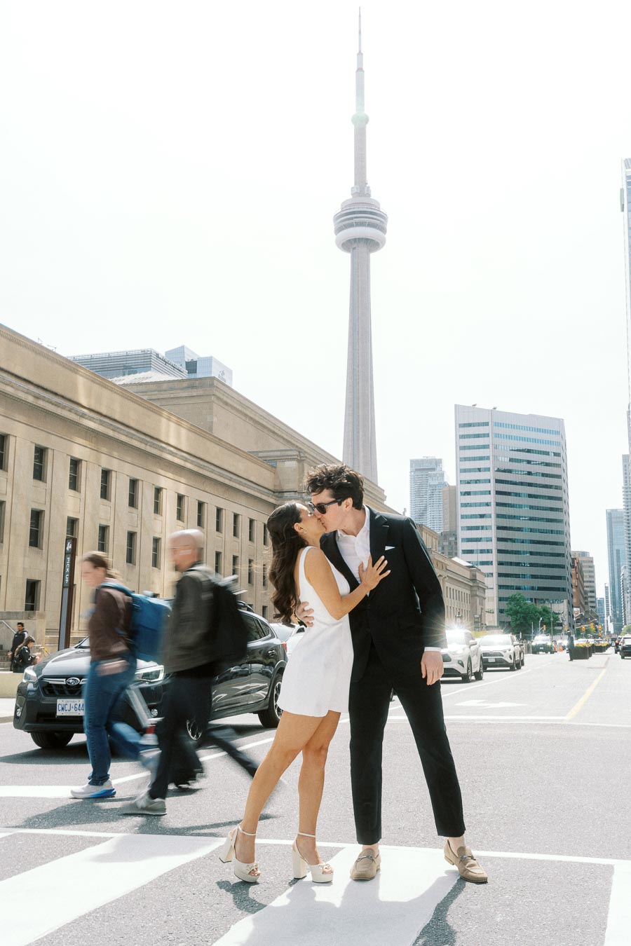 Couple kissing on a city crosswalk in downtown Toronto with Union Station and the CN Tower visible, surrounded by pedestrians and cars.