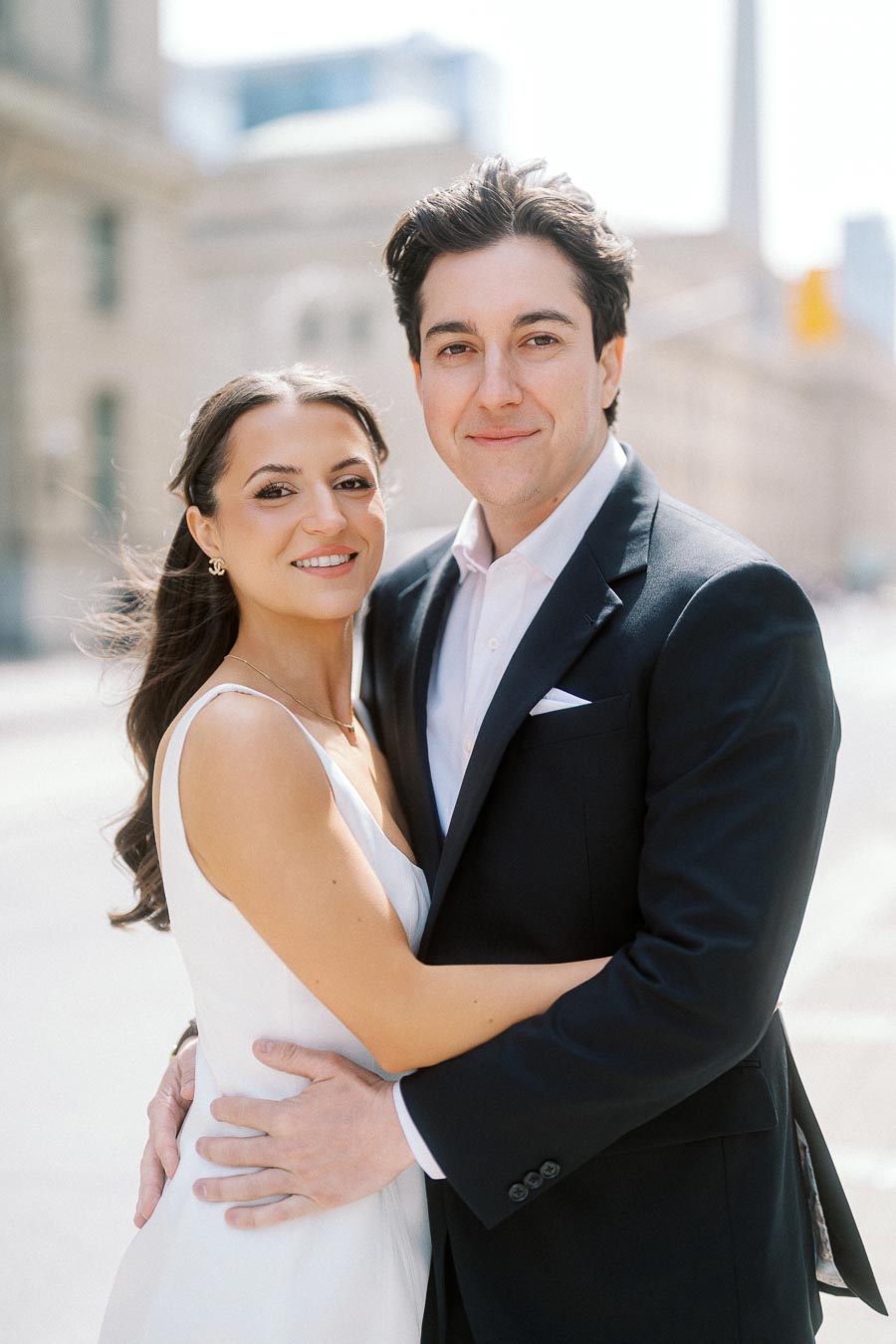 A couple stands close together on a city street, smiling at the camera, with blurred urban buildings and the CN Tower in the background.
