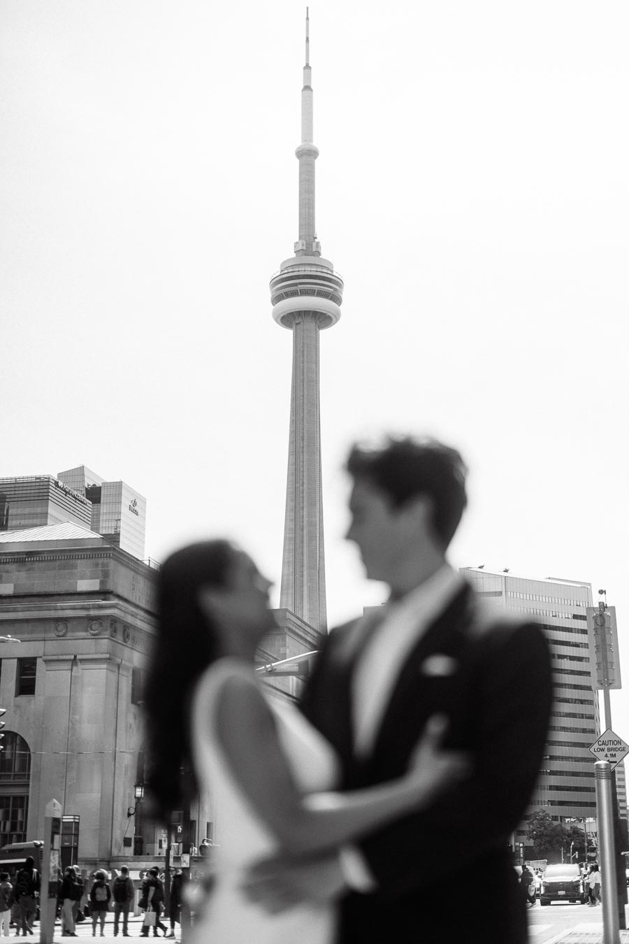 A couple in formal attire embraces out of focus in the foreground with the CN Tower and downtown Toronto buildings in sharp focus behind them.