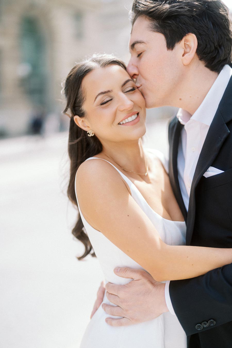 A couple embraces outdoors in soft sunlight, with the man gently kissing the woman's cheek as she smiles and closes her eyes.
