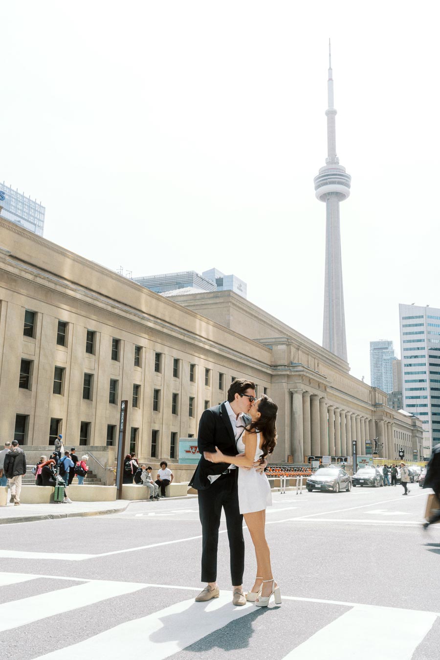 Couple kissing on a crosswalk in front of Union Station with the CN Tower visible in the background on a sunny day in Toronto.