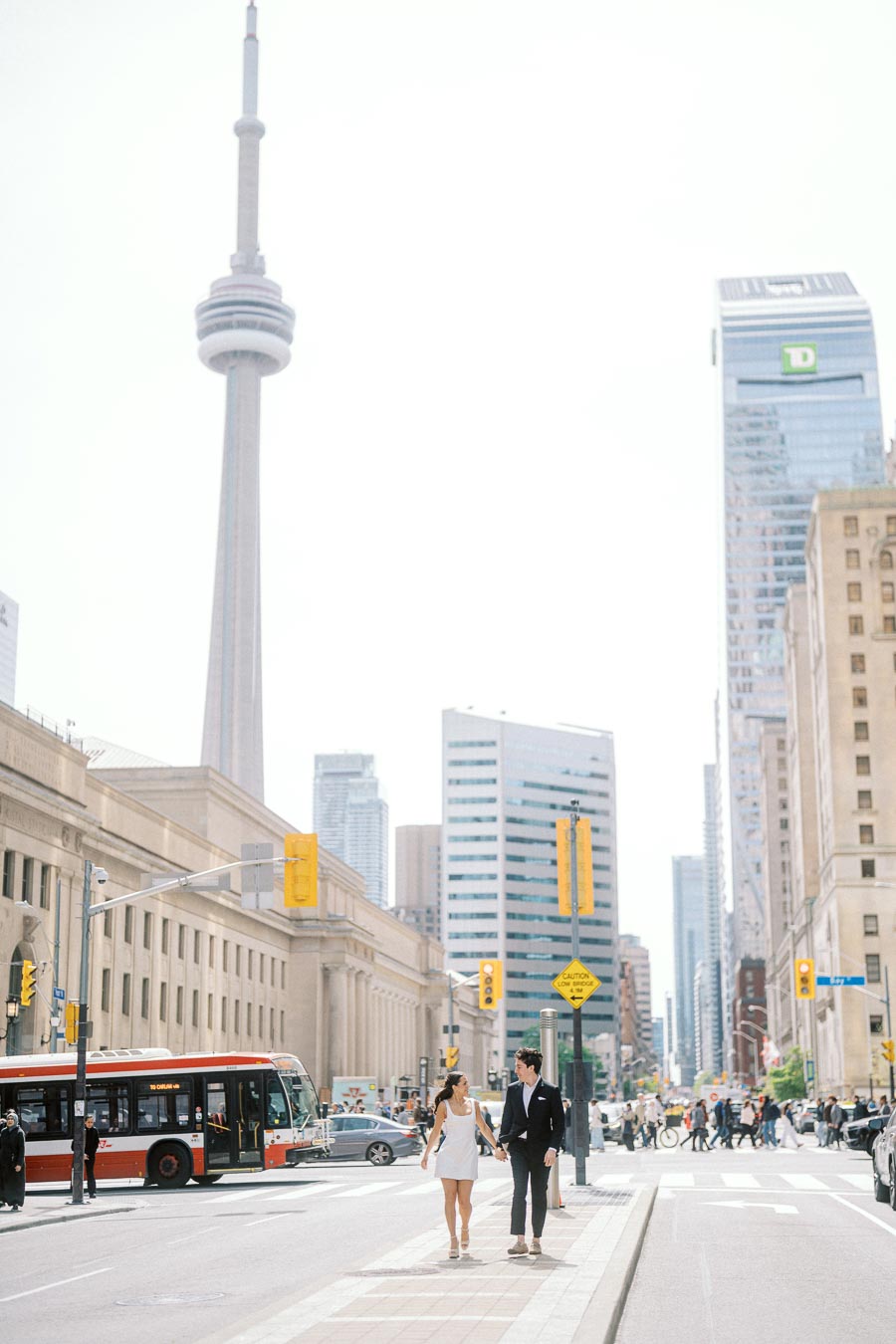 A couple walks hand in hand across a city crosswalk near Union Station in Toronto. The CN Tower and city buildings are visible behind them.