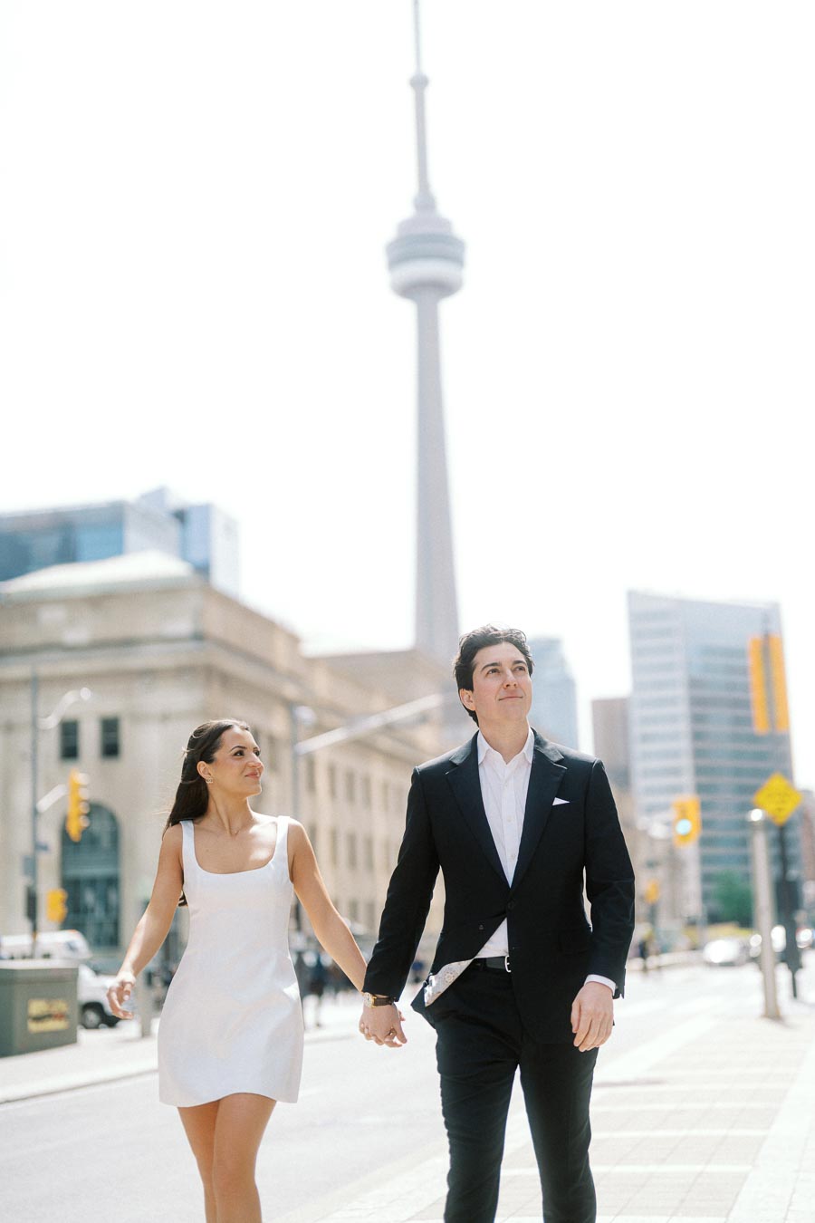 A couple dressed formally walks hand in hand on a city sidewalk in Toronto with the CN Tower in the background.