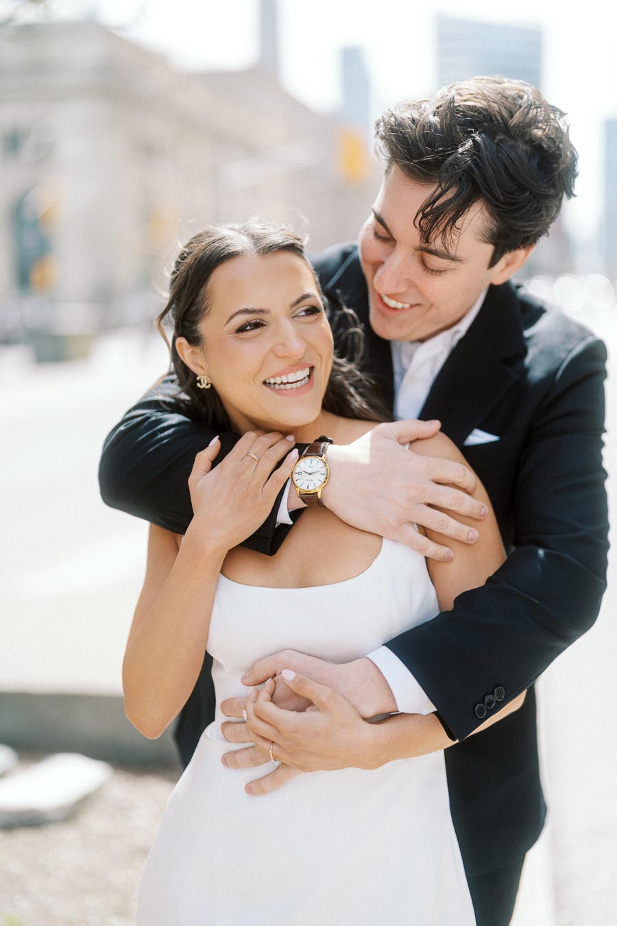 A smiling couple in formal attire embraces outdoors on a bright day in downtown Toronto, with blurred urban buildings in the background.