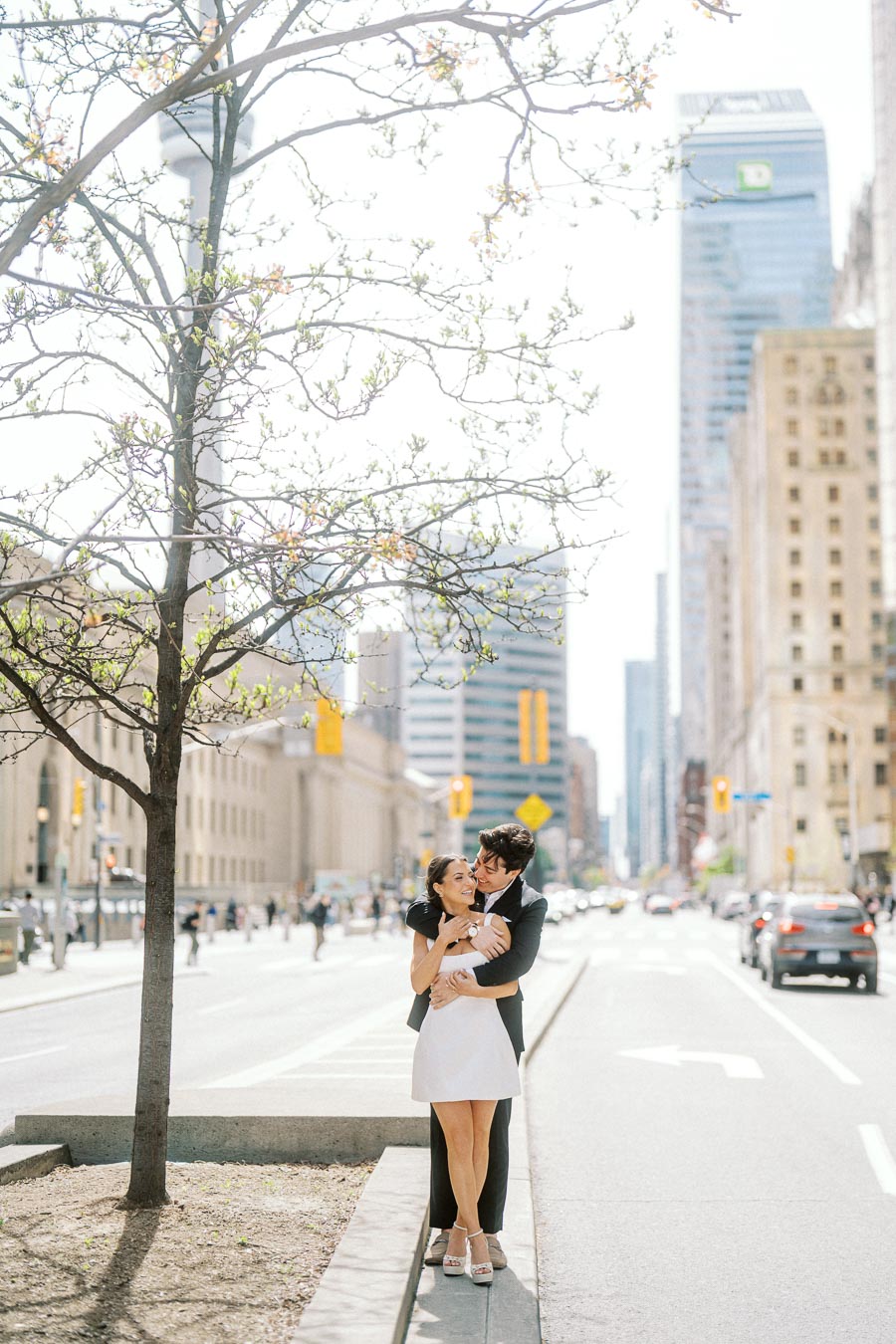 Couple embracing on a downtown Toronto street with tall buildings and a tree with budding leaves, city traffic and pedestrians in the background.