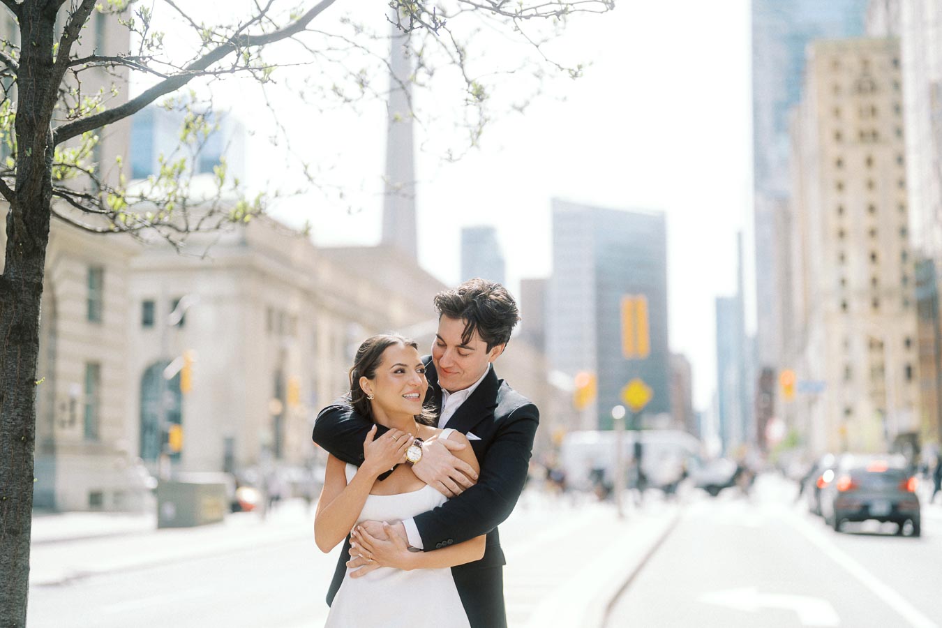 Couple in formal attire embracing on a city street in downtown Toronto with CN Tower and urban buildings visible in the background.