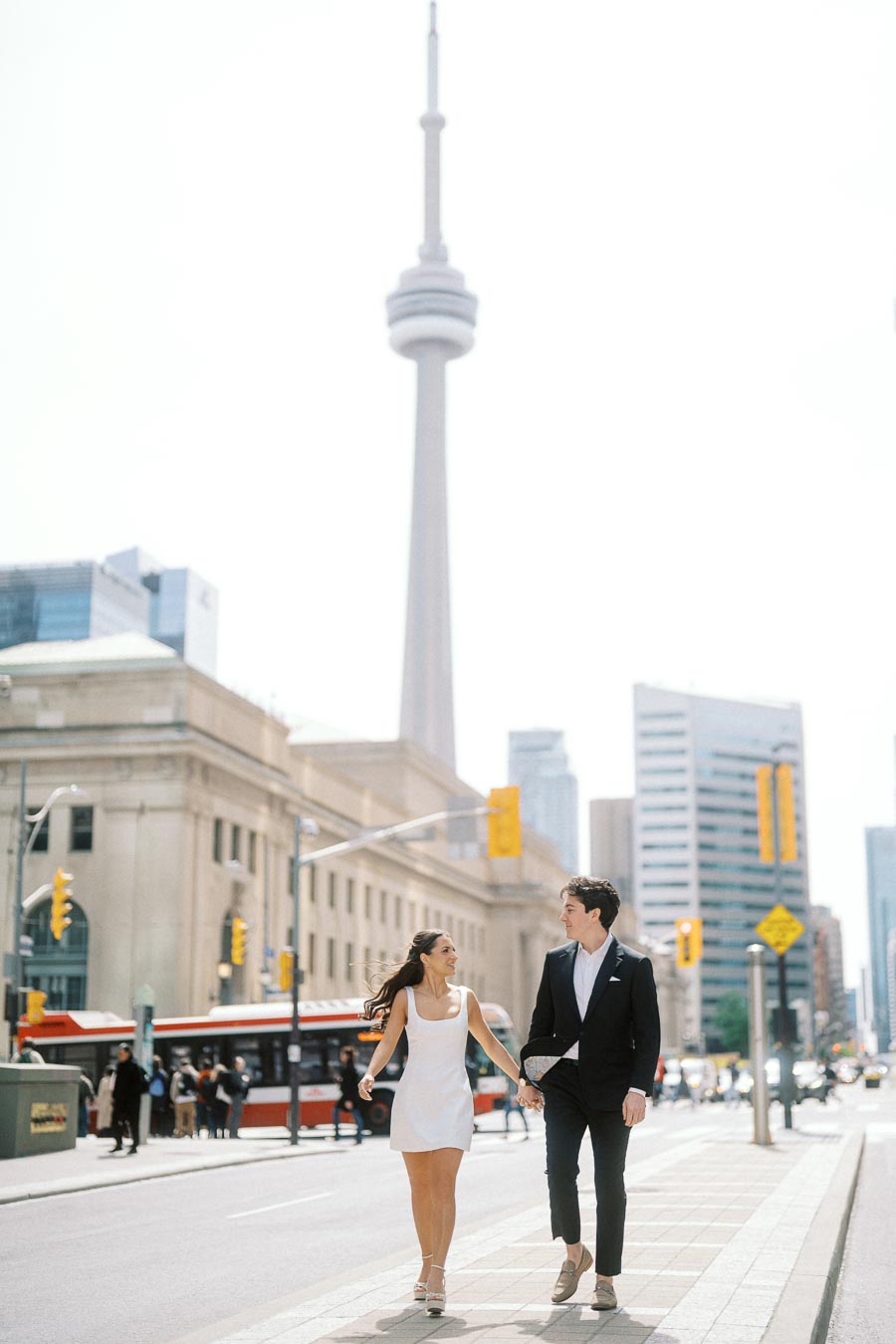 Couple holding hands and walking on a city sidewalk near Union Station with the CN Tower and urban buildings in the background.