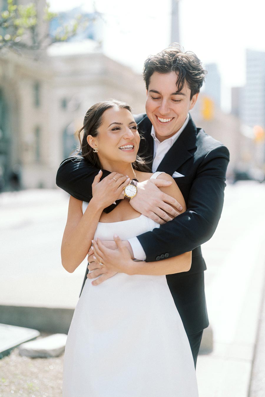 Smiling couple in formal attire embracing on a sunlit downtown Toronto street with city buildings in the background.