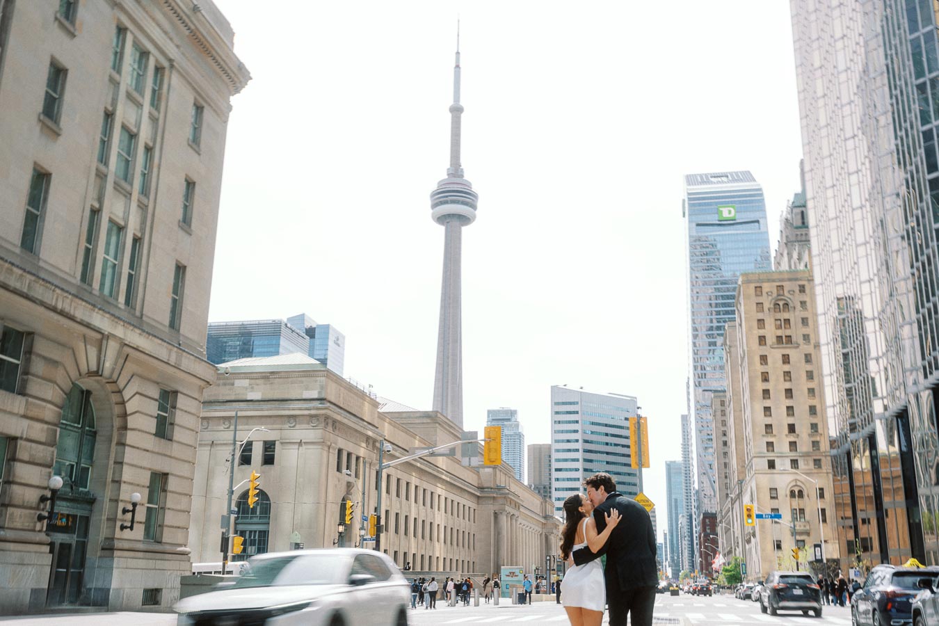 Couple embracing and kissing on a busy Toronto street with the CN Tower and urban buildings visible in the background.