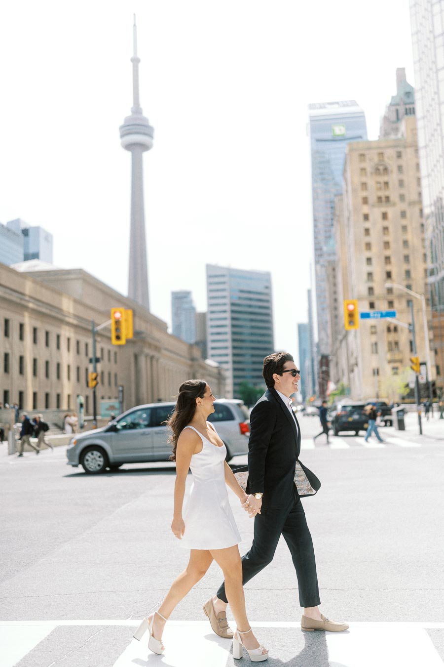 Couple holding hands walking across a crosswalk in downtown Toronto with the CN Tower and city buildings visible behind them.
