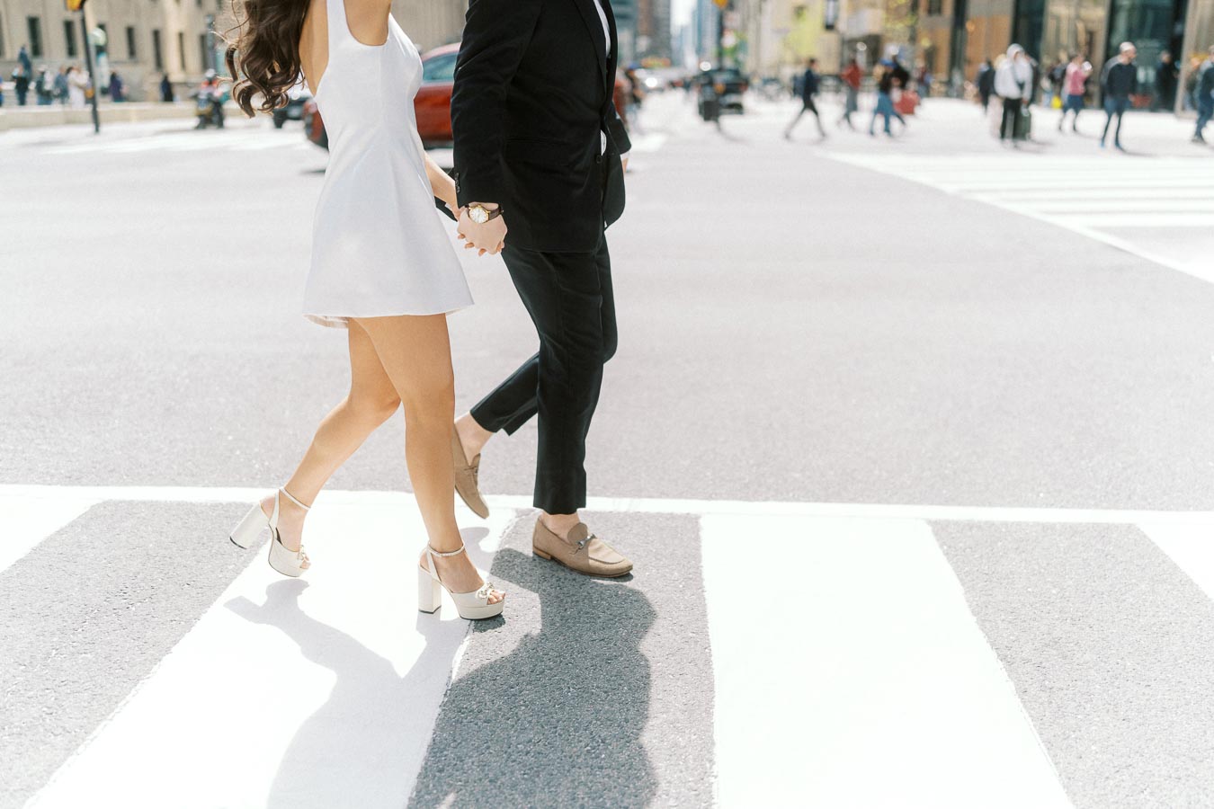 Couple holding hands and walking across a city crosswalk in downtown Toronto, dressed in formal attire, with blurred pedestrians in the background.