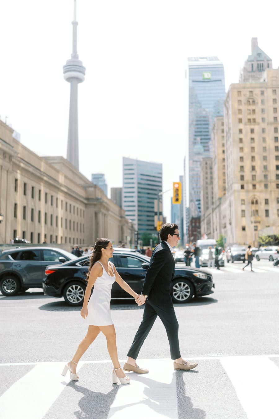 Couple holding hands and crossing a city street crosswalk in downtown Toronto with the CN Tower and tall buildings visible.