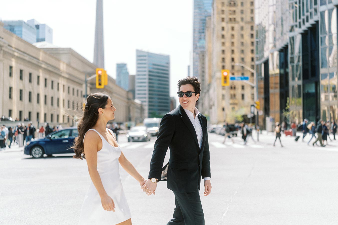 Couple holding hands and smiling while crossing a city street in downtown Toronto with Union Station and CN Tower visible in the background.