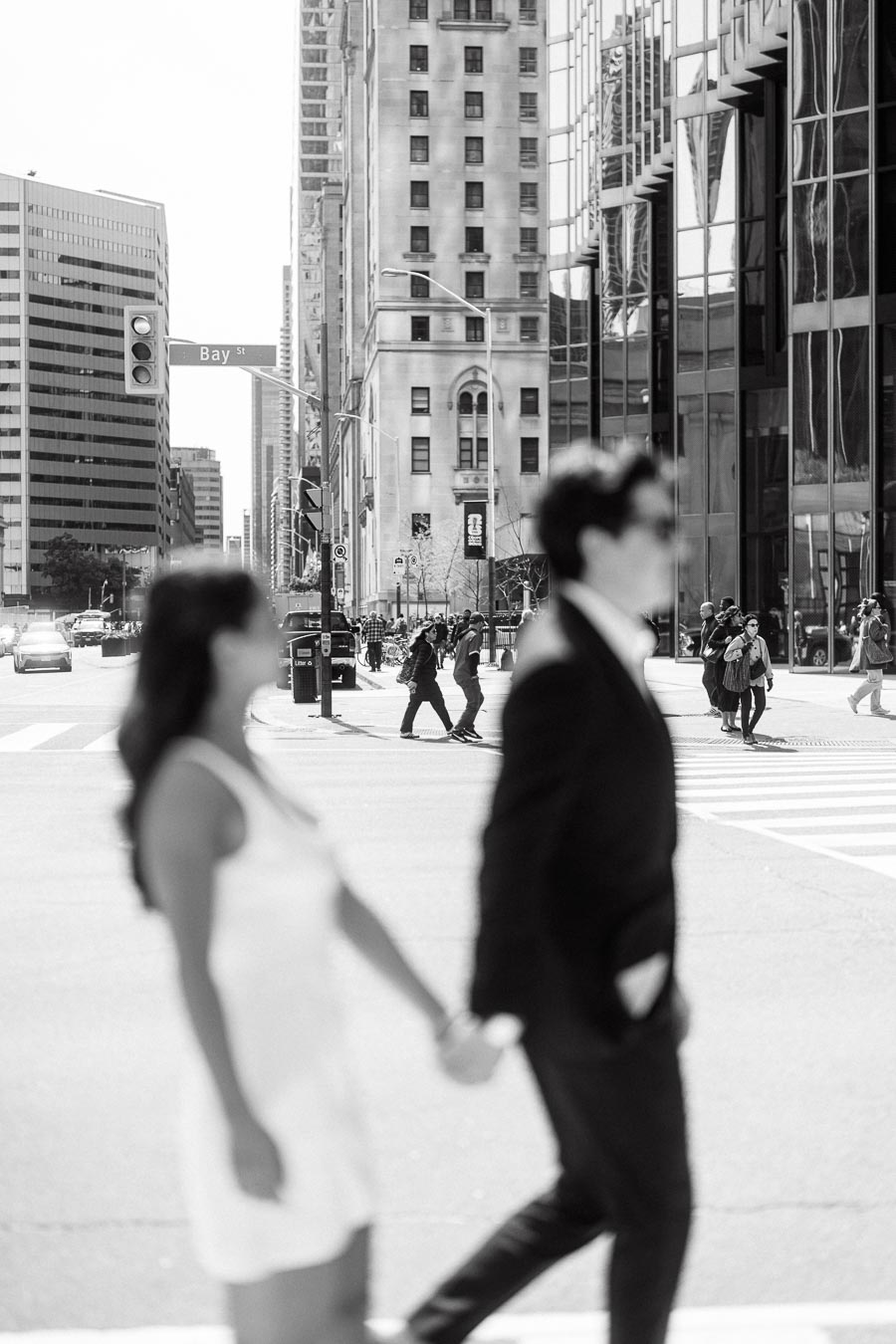 A couple holding hands and walking across a crosswalk in downtown Toronto with tall buildings and city life in the background.