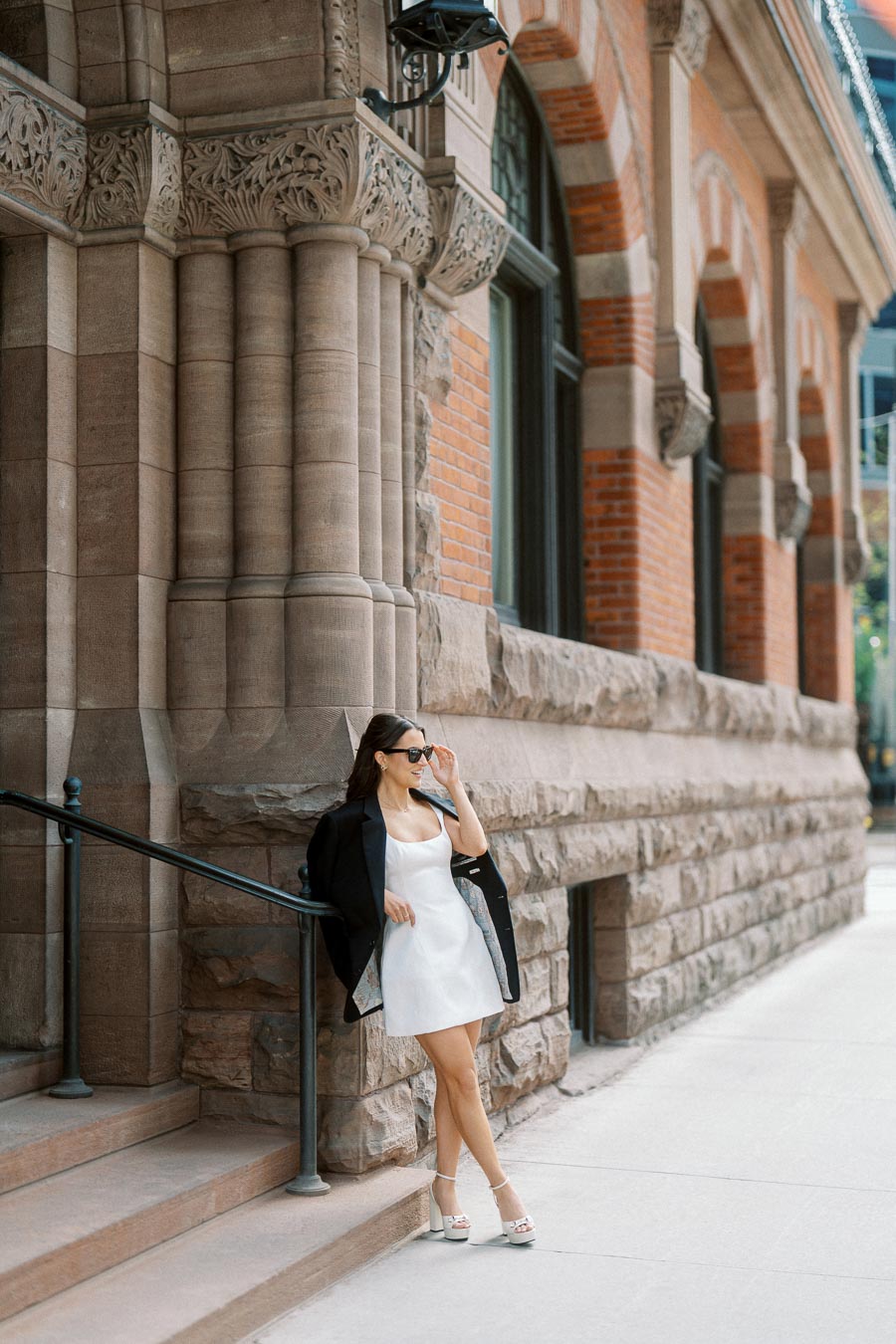 Woman in white dress and black blazer stands on city steps by ornate stone columns and brick building in downtown Ontario, Canada, late afternoon.