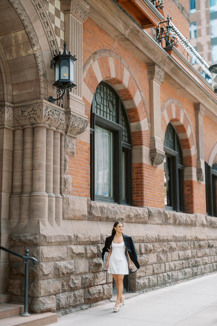 Woman in a white dress with a black jacket walking beside a historic stone and brick building with arched windows in downtown Ontario, Canada.