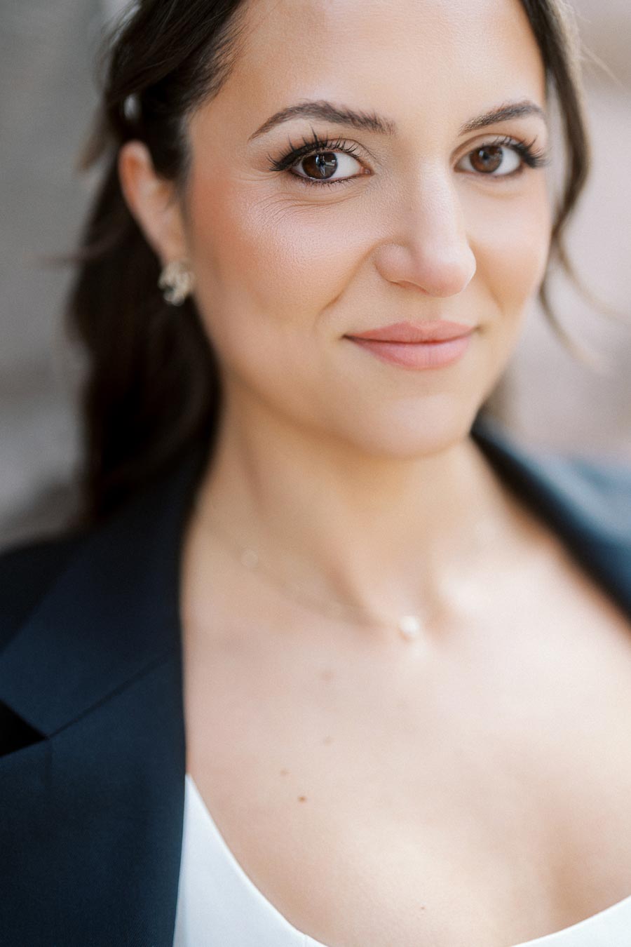 Close-up of a woman with long dark hair, natural makeup, and a navy blazer, softly smiling during an outdoor engagement photoshoot in Ontario.