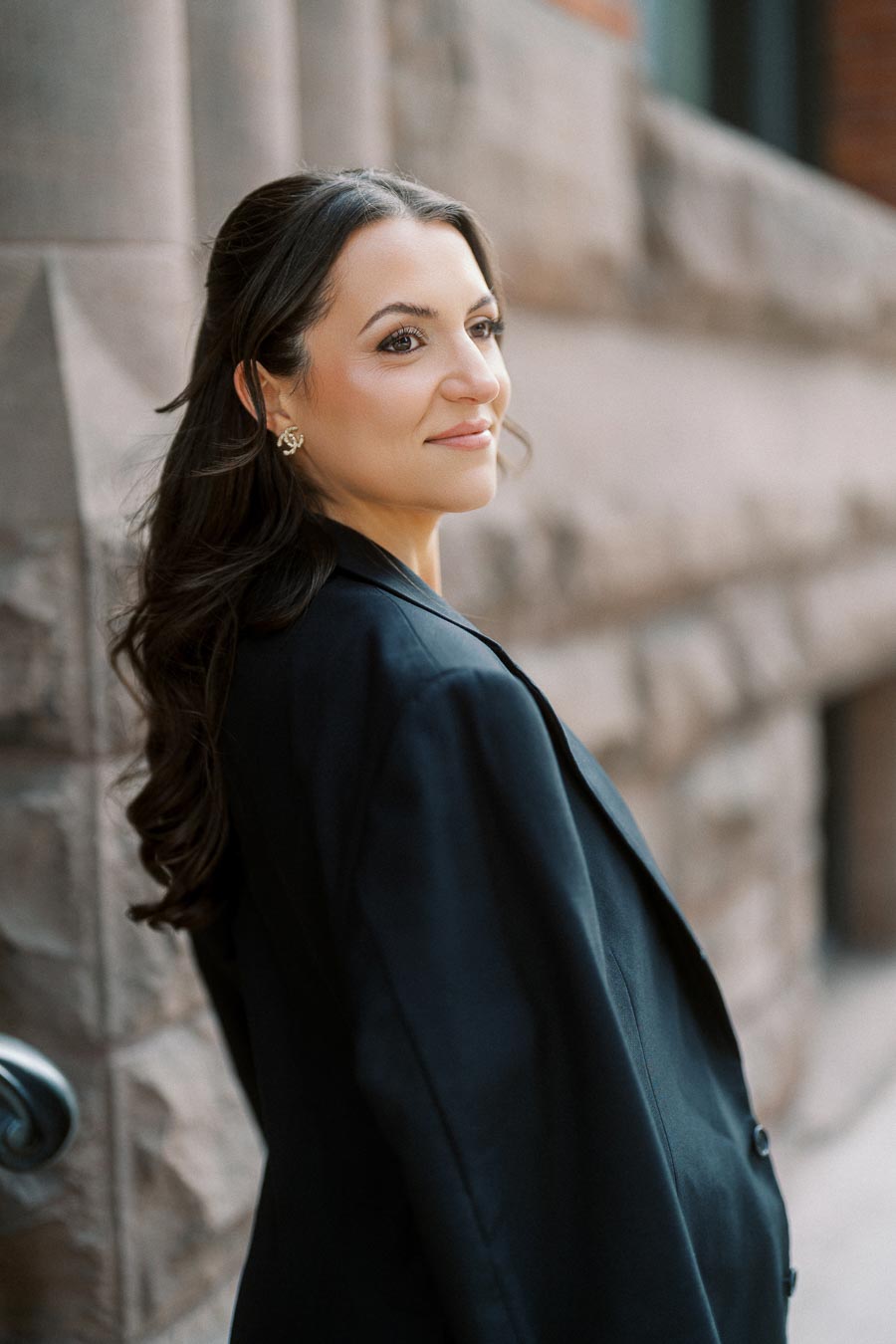 Woman with long brown hair in black blazer standing by a textured stone wall in urban Ontario, Canada, looking over her shoulder and smiling.