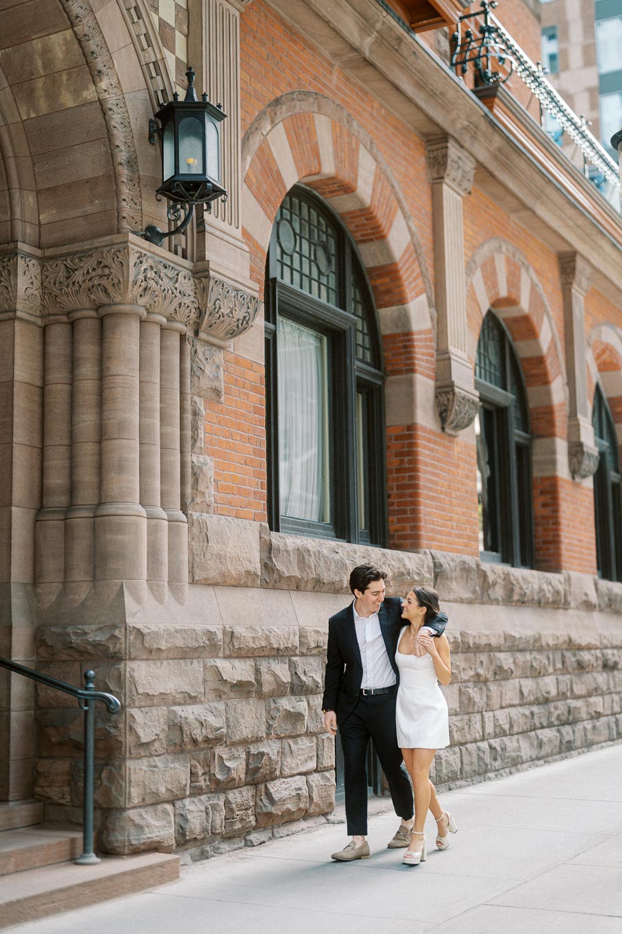 Couple walking arm in arm on a city sidewalk beside a historic stone and brick building with arched windows and ornate lantern.
