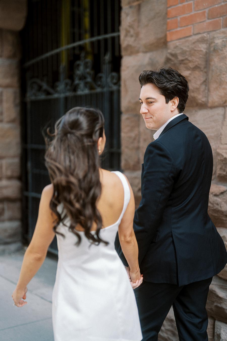 A couple dressed in formal attire holds hands while walking past a stone building in downtown Ontario, Canada, during an engagement photoshoot.