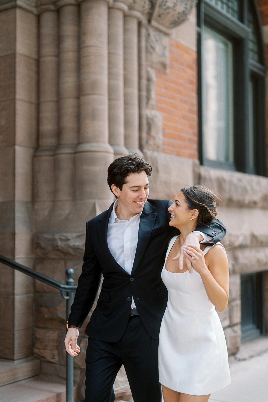 A couple in formal attire smiling and holding hands while walking outside a historic stone building with columns in Ontario, Canada.