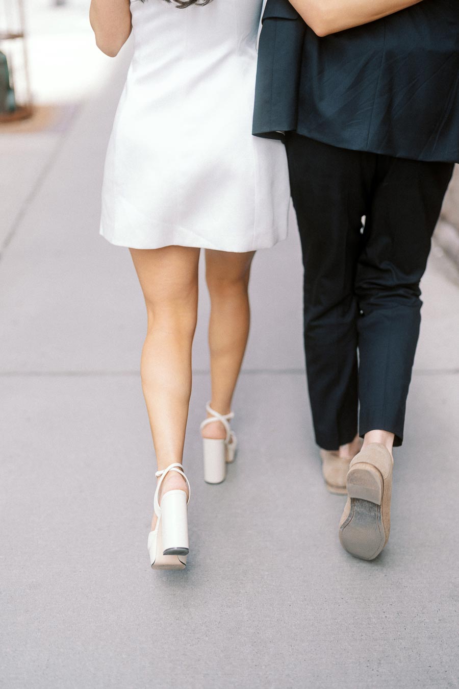 Close up of a couple walking arm in arm on a city sidewalk, the woman in a white dress and heels, the man in dark trousers and shoes.