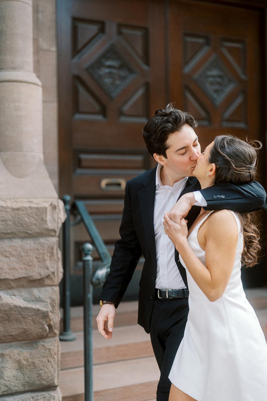 A couple in formal attire shares a kiss on stone steps in front of a large wooden door, with stone columns visible on the side.