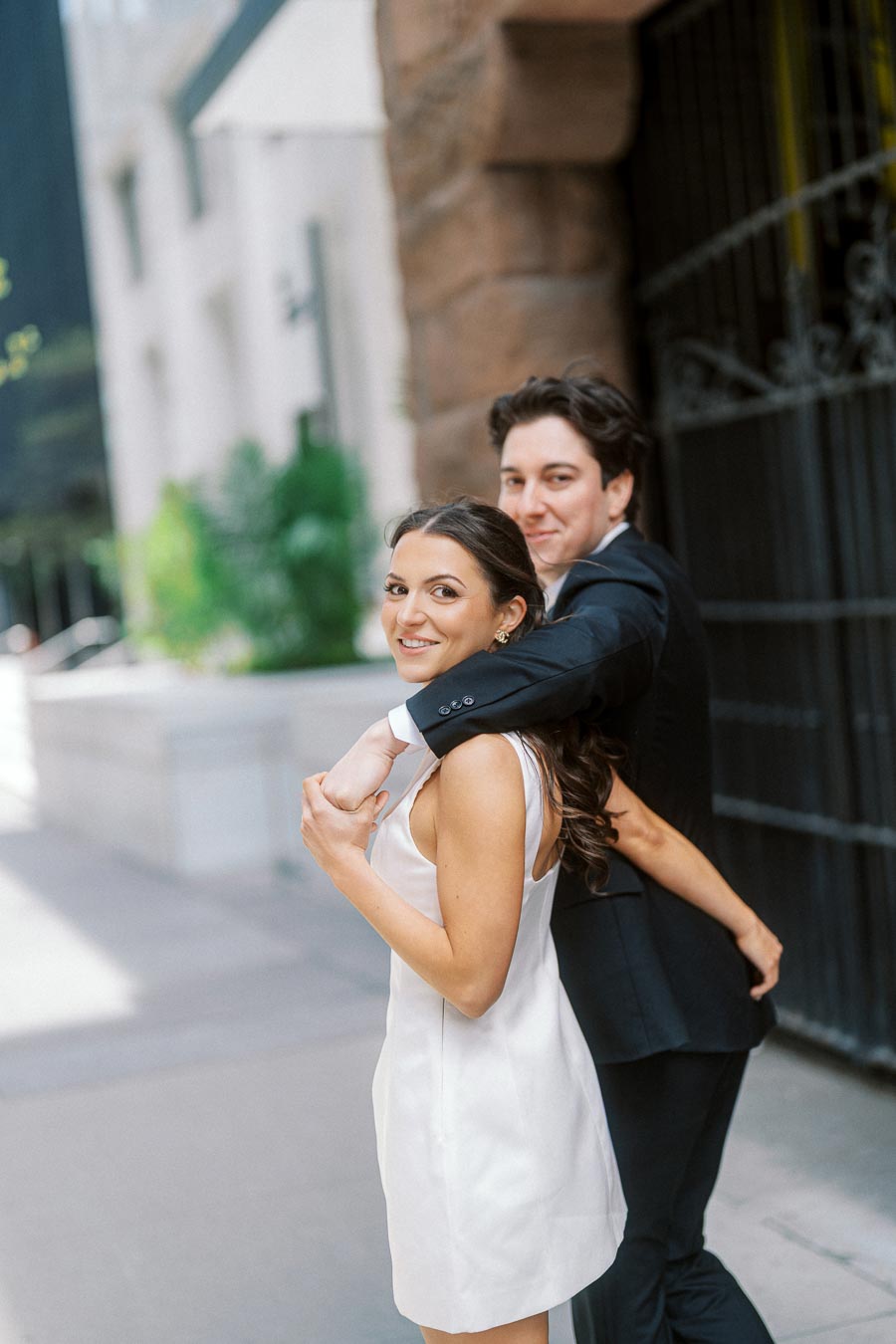 Smiling couple in formal attire embrace on a city sidewalk with stone buildings and greenery in the background.